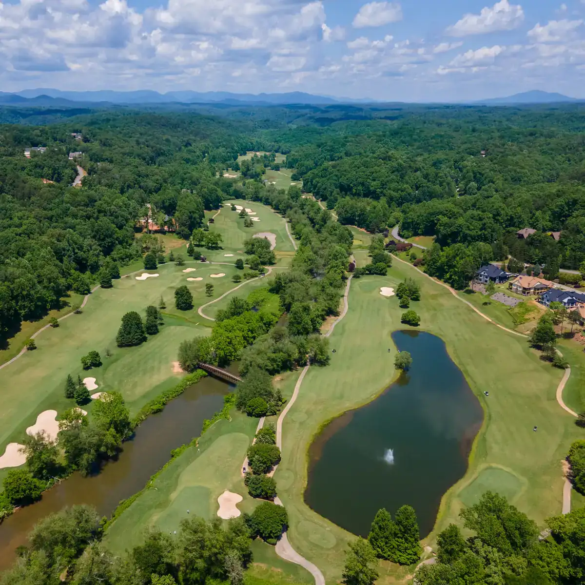 Aerial view of the Achasta golf course in Dahlonega, Georgia: fairways and sand bunkers winding through hardwood forest, a pond with a fountain, a river crossed by a footbridge, homes along the right edge, and blue ridges on the horizon.