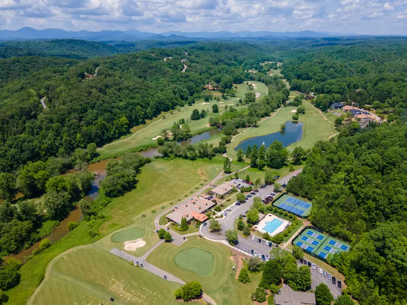 Wide aerial view of Achasta: the clubhouse, parking, tennis and pickleball courts and swimming pool in the foreground, golf fairways and ponds beyond, and forested ridges on the horizon.