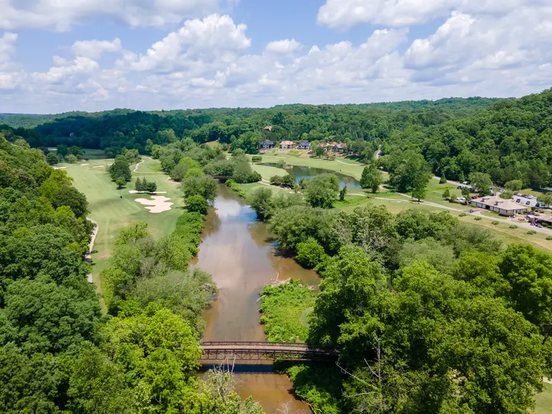 Aerial view over Achasta: a river winding between golf fairways and hardwood forest, a footbridge crossing it in the foreground, and homes on the far bank under a cloudy summer sky.