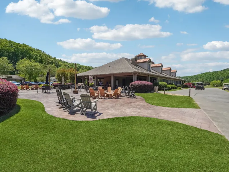 The Achasta clubhouse seen from the cart path: a stamped-concrete patio with Adirondack chairs and a fire pit in front, green lawn and wooded hills behind.
