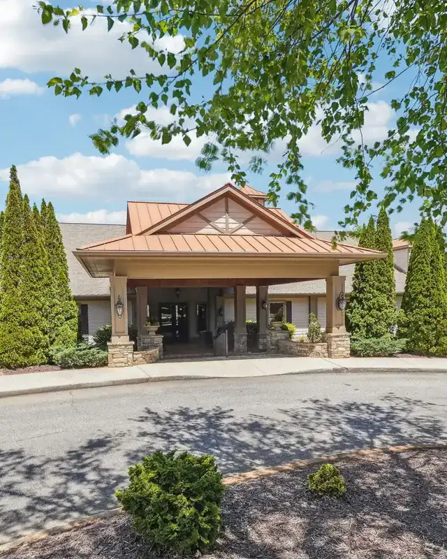 The front entrance of the Achasta clubhouse: a porte-cochere with a copper-toned gable roof on stone-based columns, tall cypress trees on either side, a curved paved drive striped with tree shadows in the foreground, and leafy branches overhead against a blue sky.