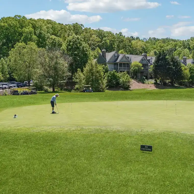A golfer putting on the Achasta practice green, with golf carts, parked cars and shingle-sided homes among the trees beyond.