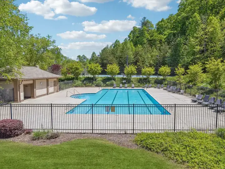 Fenced outdoor community swimming pool in Achasta with lap lanes, a wide concrete deck lined with lounge chairs, a small pool house at left, and dense green trees under a blue sky with scattered clouds.