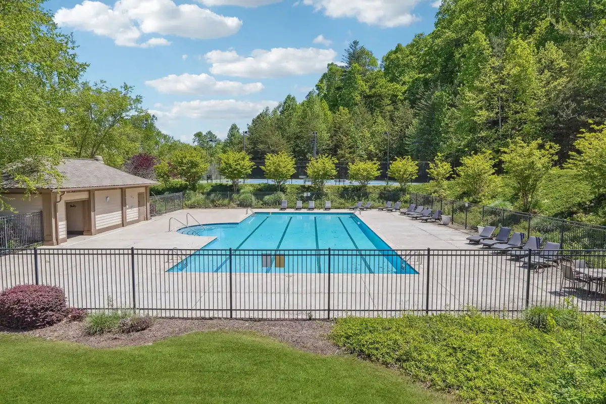 Fenced outdoor community swimming pool in Achasta with lap lanes, surrounding concrete deck, rows of lounge chairs, a small pool house, landscaped shrubs, tennis courts visible beyond the black metal fence, and dense green trees under a blue sky with scattered clouds.