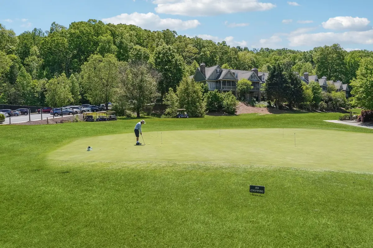 A golfer putting on the Achasta practice green, with golf carts, parked cars and shingle-sided homes among the trees beyond.